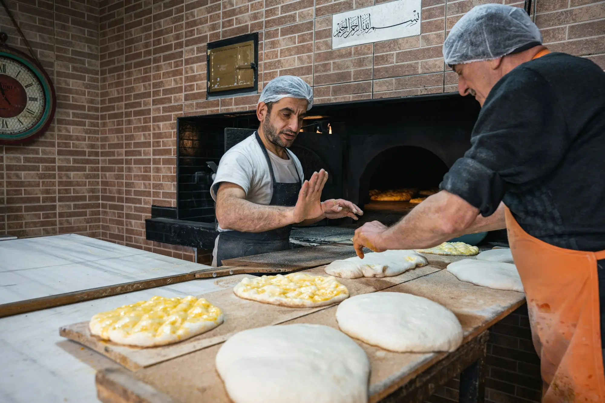 Bakers shaping and preparing dough on a floured bench in front of a brick oven in a small bakery.