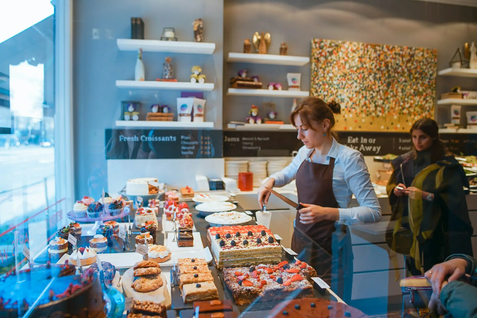 A staff member behind a bakery counter serving cake and pastries to a customer.