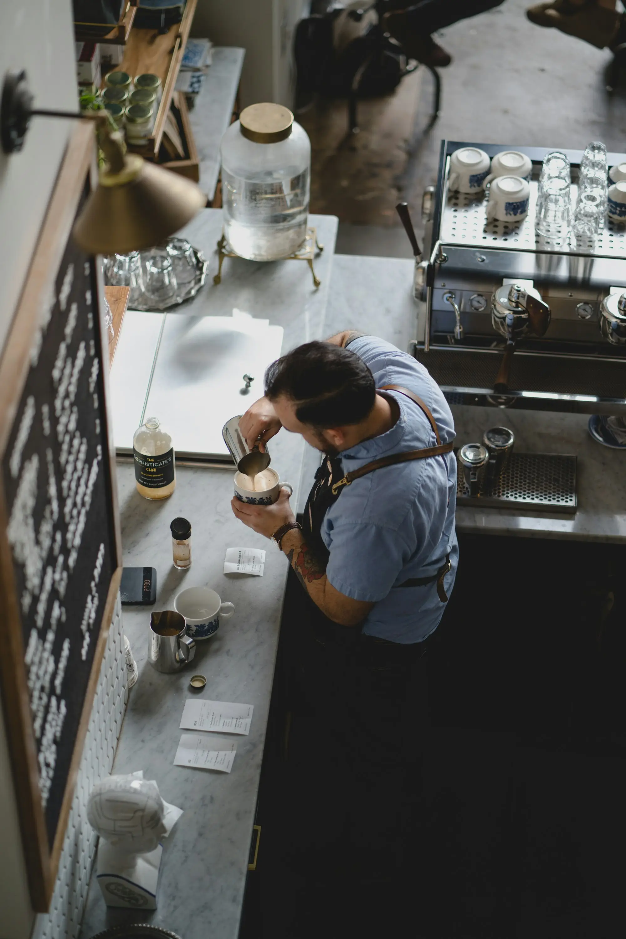 A cafe owner in a blue shirt pouring coffee at the espresso machine behind the counter.