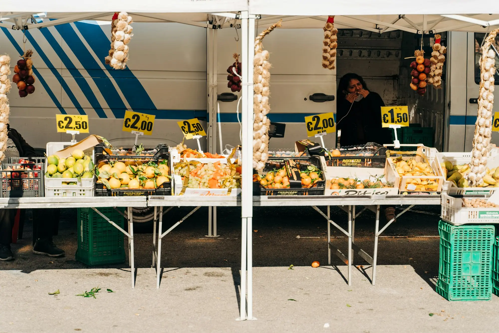 A stallholder arranging fresh fruit on a market stall table with hand-written price signs.