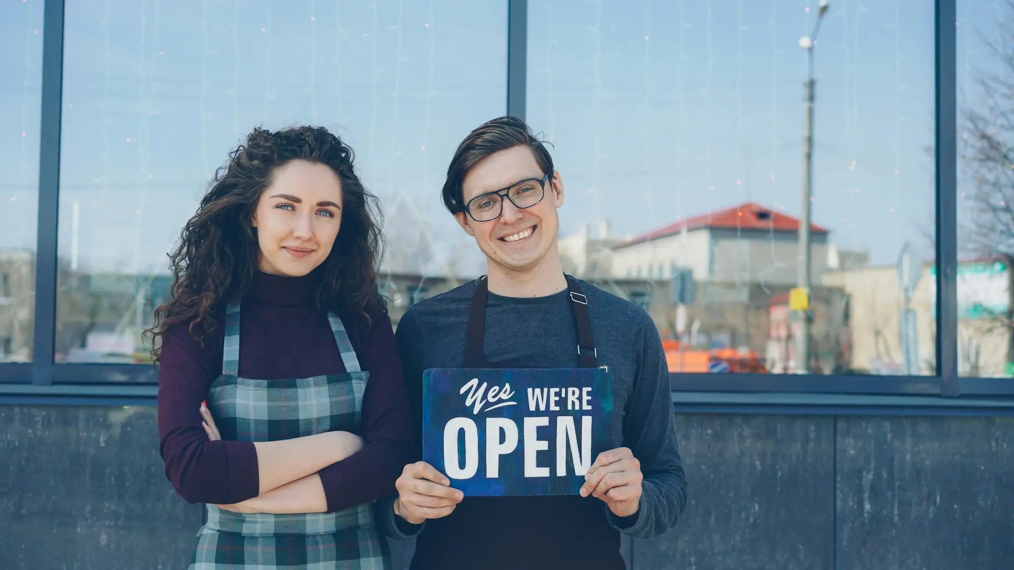 Two small shop owners standing at their front window holding up an open sign.