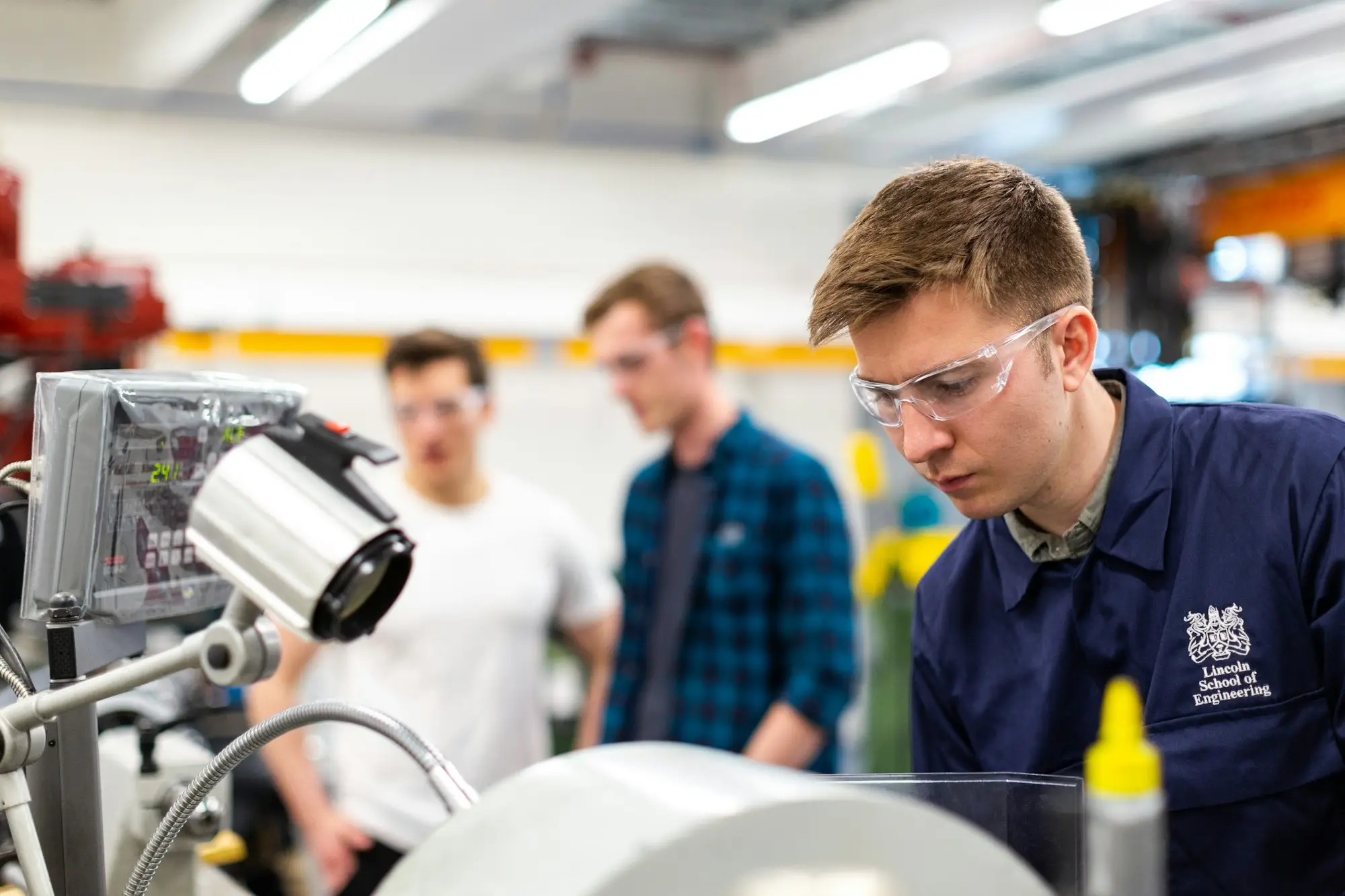 A young apprentice in safety glasses learning a machine task in a small workshop while colleagues look on.