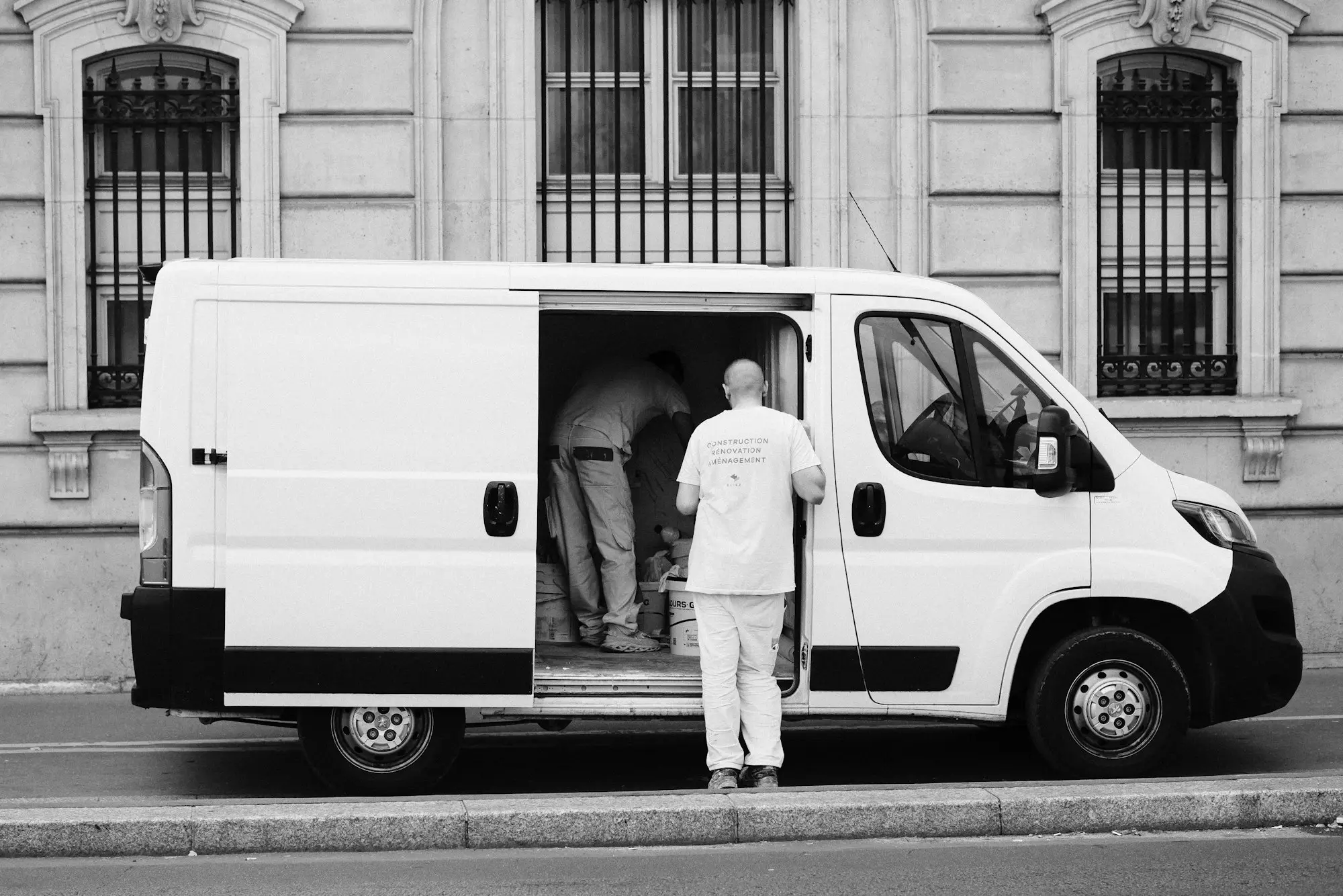 A tradesman in work clothes standing at the open side door of a white work van, loading up for the job.